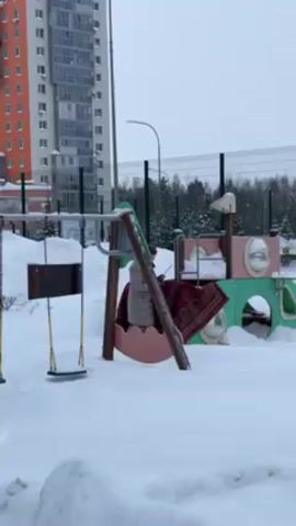 A woman is shaking out a large patterned rug on a snow-covered playground. She is wearing a winter coat and hat. The playground includes a swing set and a play structure with tunnels and windows. Behind the playground is a tall, modern apartment building, a black metal fence, trees, and a snowy landscape. The sky is overcast. The snow is deep.