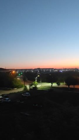This is a vertical, slightly shaky, wide-angle video captured at dusk from a high vantage point looking down onto a street scene. The sky is a gradient of deep blue at the top transitioning to orange and pink near the horizon. In the distance, there is a town or city with buildings and streetlights illuminated. A street runs horizontally across the middle of the frame, with cars parked along its side and a single person walking on the sidewalk. Trees and bushes line the street, and some are visible in the foreground. The overall mood is calm and peaceful, with the transition from day to night clearly visible.
