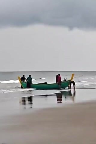 A group of five people are seen interacting with a teal-colored boat with yellow accents on its bow and stern. The boat is partially submerged in shallow water at the edge of a beach. The sky is overcast with gray clouds. The water is calm with small waves. The beach is wet and reflective. The people appear to be either pushing or pulling the boat. Their actions suggest they are preparing to launch or land the boat.