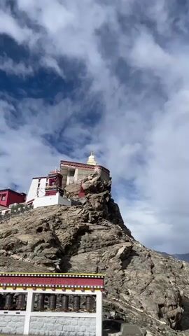 A vertical video shot in the daytime shows a rocky mountain with a monastery on top. In the foreground, prayer wheels are visible. The video pans to the right, revealing a valley with cultivated fields, trees, and distant mountains under a cloudy sky.