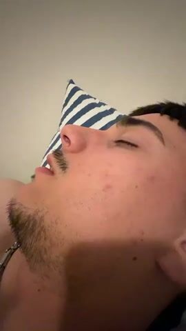Here's a description of the video:

A close-up shot depicts a young man lying on his back with eyes closed and a slight stubble beard. He rests on a pillow with blue and white stripes. A hand enters the frame, touching the man's nose and cheek with painted nails. The man appears to be asleep or resting undisturbed by the touch. The background is a plain, light-colored wall.