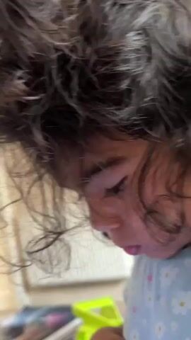 A close-up shot of a young child's head. The child has dark, curly hair that obscures most of their face, but their profile is visible. They are looking downwards intently, with their eyes closed. The child is wearing a light blue t-shirt with a pattern of white flowers. The background is blurred, but suggests an indoor setting with some colorful objects, possibly toys or books, on the floor. The lighting is soft, with some light catching the child's hair and cheek.