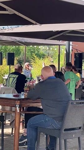 This video captures an outdoor scene at a restaurant or cafe in Saint-Just-Saint-Rambert, France, during a bright, sunny day. Several individuals are seated at wooden tables under large dark umbrellas, enjoying a live music performance.

In the mid-ground, a woman with blonde hair, dressed in a light denim jacket over a dark top, is standing and singing into a microphone. Two large black speakers on stands are positioned behind her, indicating a professional setup for the performance.

In the foreground, a man with short dark hair, wearing a black t-shirt with a green and black checkered pattern and the word "WRITE" prominently displayed, is seated at a wooden table. To his right, a bald man in a dark grey long-sleeved shirt and blue jeans is seated with his back to the camera, looking towards the performer. Both appear to be enjoying the music.

Other patrons are scattered around, either seated at tables or standing. A young boy in a green t-shirt is visible standing near a table on the right side of the frame. Drinks, possibly cocktails, are visible on the tables. The number "40" is marked on the edge of the wooden table in the foreground.

The background features lush green foliage, suggesting trees and bushes, under a clear sky. The atmosphere appears relaxed and pleasant, with people enjoying the outdoor setting and the live entertainment.