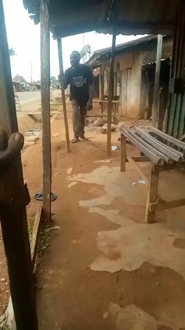 A man in a black t-shirt and brown pants walks into the frame and sits on a wooden bench. The bench is covered in what appears to be bamboo poles. The man is standing in what looks like an outdoor market stall, with dirt ground and corrugated metal siding on the right side of the frame.