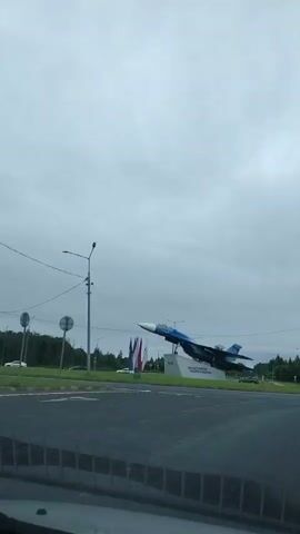 A low-angle, stationary shot looking out from a vehicle toward an outdoor monument. The monument features a blue and white fighter jet mounted on a grey pedestal with text. A pole with a light fixture and a traffic sign are visible to the left of the jet. Several flags, appearing to be red, white, and blue, are planted in the grass behind the jet. The sky is overcast and grey. The foreground shows the dark interior of the vehicle, including the windshield wipers. The scene appears to be a roundabout or intersection.