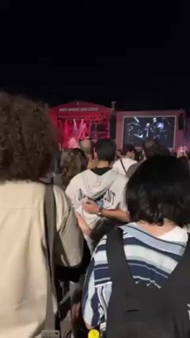 Here is a detailed description of the video:

This video shows a concert in an outdoor venue at night. The camera's point of view is from the crowd, looking towards a stage in the distance. The stage is elevated and has a red hue with bright lights shining on it. There are multiple screens showing the performers in black and white. People are standing in the foreground with their backs to the camera. The crowd is dense, with people of varying heights and hairstyles. Some people are wearing casual clothing like hoodies, jackets, and backpacks. One person is waving what appears to be a red and white Turkish flag. The overall atmosphere seems to be lively with the sound of music and cheering in the background.