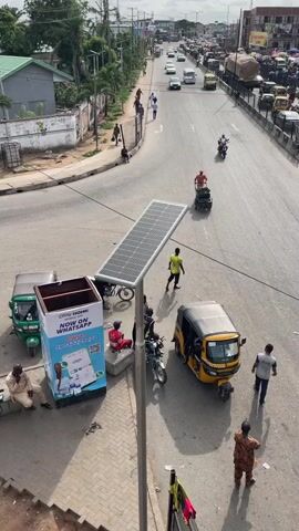 The scene is a sunny, high-angle daytime shot of a busy intersection in what appears to be a city in Nigeria. Cars and tricycles (auto rickshaws) move along the main road, which has two lanes in each direction. Several pedestrians are visible.

In the foreground, on a paved section of the sidewalk, a solar panel is mounted on a pole. Next to the solar panel is a promotional display for “OKAY HOME” with the tagline "NOW ON WHATSAPP" and a phone number. Several people are gathered around this area. One man is sitting on a low concrete barrier. Another is standing nearby talking with someone who is partially obscured. A man in a red shirt is sitting on a low wall. Two motorbikes are parked near the solar panel.

Across the intersection, in the direction of the flow of traffic, a man is pushing a loaded handcart. Other people are walking along the side of the road. The background shows buildings and trees along the street, and additional traffic further down the road.