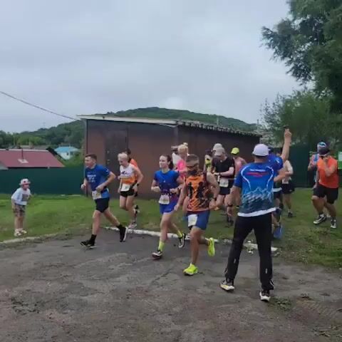 A group of runners, both male and female, are participating in an outdoor race. They are running on a dirt path. Many of the runners are wearing athletic clothing and bib numbers. The environment appears to be a rural or semi-rural area with some buildings and trees in the background. The sky is overcast.