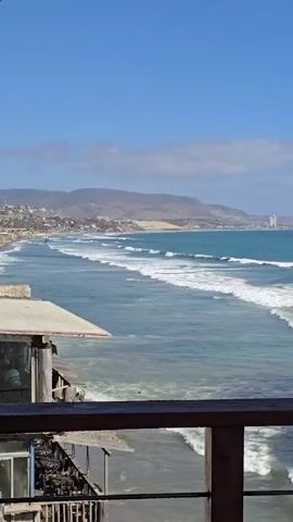The video captures a scenic view of a beach with turquoise water and rolling waves. In the foreground, a wooden railing and parts of a rustic building are visible. The building appears weathered and has a simple design, with some wooden beams and what looks like a corrugated roof. Beyond the building, the sandy beach stretches along the coastline, meeting the ocean. Gentle waves are breaking on the shore, creating white foam. In the distance, a line of hills forms the backdrop, with some structures and buildings scattered along the hillside and a taller building visible further down the coast. The sky is clear and blue, with a few wispy clouds. The overall impression is a relaxed, coastal environment.