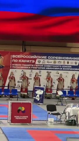 A choir of seven women in traditional clothing is singing a capella while standing in front of a banner in Russian. The banner is for the All-Russian Karate Competition, a memorial cup dedicated to a Soviet hero, Grigory Ivanovich Boyarinov. The women are holding microphones and standing behind a row of blue chairs. To the right of the women, a man in a military uniform is saluting. A camera crew is filming the scene. A World Karate Federation sign is visible in the foreground. The floor is red and blue.