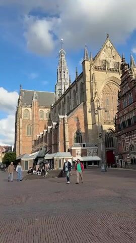 The video displays a wide-angle view of a cobblestone plaza in front of a large, ornate brick building with a tall spire. The building appears to be a cathedral or church with Gothic architectural elements, including arched windows and flying buttresses. The sky is blue with scattered white clouds, suggesting daytime. Several people are walking across the plaza. In the foreground, three individuals are walking from left to right. One person is wearing a light-colored top and dark pants. Another person, in the middle, is wearing a blue jacket and dark pants. The third person is wearing a red top and khaki pants. Further in the background, more people are scattered throughout the plaza, some sitting near a statue. The overall scene is a bustling public square with historical architecture.