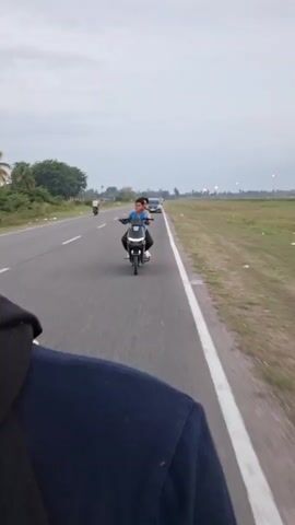 The video shows a road from the perspective of someone on a motorcycle, with another motorcycle approaching in the opposite direction. On the approaching motorcycle are two people. The person driving the motorcycle appears to be older than the person sitting in front. They are both wearing shirts and appear to be smiling. In the background of the shot there are vehicles including cars and a motorcycle. There are also various trees and foliage, and a grassy field. The sky is overcast.