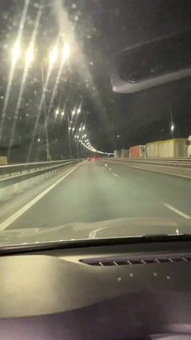 The video shows a first-person view from inside a car driving on a highway at night. The windshield is visible in the foreground, with some reflections or glare. The road ahead is well-lit by numerous streetlights extending into the distance. Several bright streetlights are visible on the left side, illuminating the highway. In the far distance, the red taillights of other vehicles can be seen. To the right of the highway, large structures resembling containers or trucks are visible. The car appears to be moving at a steady pace. No people or visible text are present in the frame.