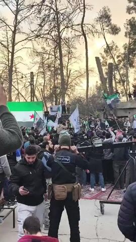 The video shows a large crowd of people gathered outdoors. Many are waving flags with the colors green, white, and black, with red stars, likely Syrian flags. Some people are standing on elevated platforms, addressing the crowd. There are also individuals in uniform with "قيادة الأمن الداخلي" written on their backs, which translates to "Internal Security Leadership." A person in the foreground is looking at their phone. The environment appears to be a park-like area with tall trees.
