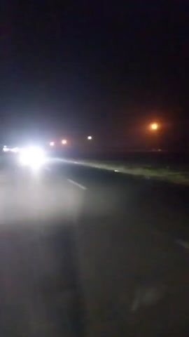 A nighttime view shows a road from the perspective of someone driving or riding. Streetlights line the right side of the road. In the distance, car headlights approach.