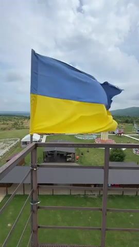 A Ukrainian flag flutters on a metal pole attached to a railing. The flag has two horizontal stripes, the top one blue and the bottom one yellow. Behind the flag, a grassy area is visible with several buildings, including a long, dark-roofed structure. In the distance, there are rolling hills and a cloudy sky. The camera is positioned on a platform or tower looking down at the scene.