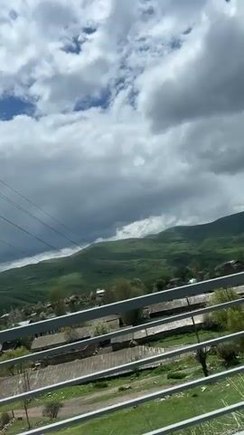 The video shows a landscape view from what appears to be an elevated position, possibly a balcony or bridge. The foreground features a white railing. Beyond the railing, there's a view of a village or rural area, with houses and buildings visible among greenery. In the distance, rolling green hills or mountains are visible, topped by a sky with scattered clouds. There are power lines visible in the upper left portion of the frame. There is no evident human activity, but a voice can be heard.