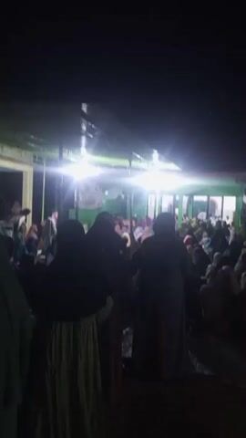 A group of people, mostly women wearing headscarves, are gathered outdoors at night under a canopy. Bright lights illuminate the scene, casting shadows and creating a somewhat hazy atmosphere. The people appear to be seated on the ground or on low seating. Some individuals are standing and interacting. The background shows a building with windows. The overall impression is of a community gathering or event taking place after dark.