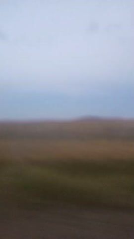 The video shows a motion blur of a landscape. The sky is a pale blue with some wispy clouds. The horizon is a gently rolling hill, possibly covered in grass or other low vegetation. The foreground is also blurred, suggesting movement, possibly from a vehicle. The colors are muted, indicative of either dawn, dusk, or an overcast day. There are no discernible people or specific objects.