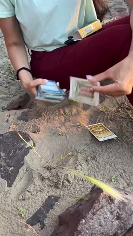 A person with dark hair, wearing a light blue shirt and maroon pants, sits on sandy ground. They are holding a deck of tarot cards in their left hand and are in the process of drawing a card with their right hand. Two tarot cards are already laid out on the sand in front of them. The environment appears to be a beach or sandy area with sparse vegetation and rocks. The video then shifts to a wider shot of a beach scene at sunset, with the sun low on the horizon over a body of water. The sky is filled with orange and yellow clouds. A young girl with long brown hair, wearing a white t-shirt, is partially visible in the foreground on the right side of the frame.