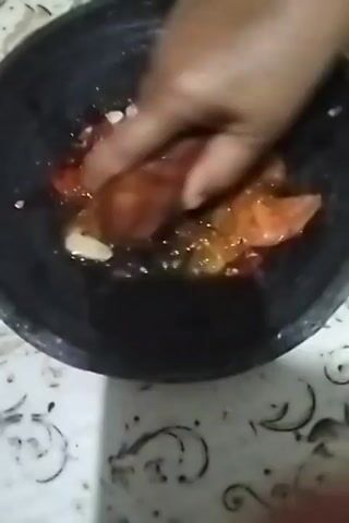 A hand is seen mashing tomatoes and garlic in a dark mortar with a pestle. The mortar is black and circular, and the ingredients are red and white. The hand is moving vigorously, crushing the ingredients. The background is a patterned surface, possibly a table cloth or countertop. The video is a close-up shot of the mortar and pestle.