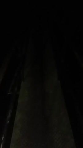 The video depicts a dark, enclosed environment, likely at night or in an unlit space. The primary visible elements are a textured, dark gray surface, possibly a walkway or bridge, and dark water. The surface features a pattern resembling diamond plate or grates, extending forward and appearing to be a path. On the left side, a dark structure or wall runs parallel to the path. Towards the end of the video, dark water with visible splashes and reflections of light becomes more prominent on the right side. The camera moves forward, suggesting a journey through a tunnel, underpass, or along a canal. The sound of water splashing is audible. No people or discernible text are visible.