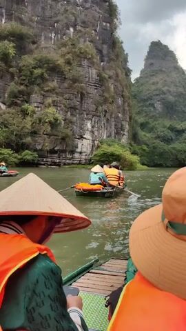 The video shows a group of people rowing boats through a river canyon. They wear orange life vests, and some wear hats, including traditional conical hats. The boats are narrow and low in the water. The river flows between tall, rocky cliffs covered in green vegetation. The sky is overcast. Other boats with people are visible ahead, and a red flag can be seen on one of the boats. The camera follows behind one of the boats.
