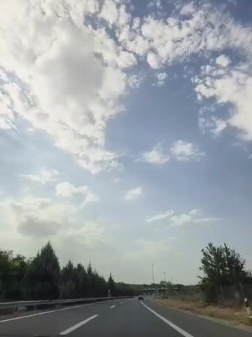 The video shows a view from inside a vehicle traveling on a highway. The sky is blue with scattered white clouds. On the left side of the road, there is a line of trees. There are also traffic signs visible, including speed limit signs. The road is paved and has lane markings. In the distance, there appears to be a toll booth or an exit. The overall environment is that of a highway drive on a sunny day.