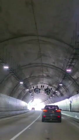 A dark, concrete tunnel with a bright opening at the end. A dark-colored SUV is driving down the road within the tunnel, and another car is visible in the distance ahead. Lights are mounted on the ceiling of the tunnel at intervals, and there are large ventilation fans also mounted on the ceiling. The tunnel walls are visible, some of them lined with white material.