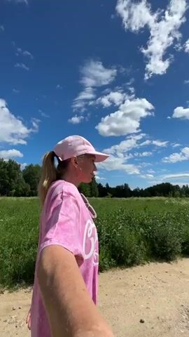 A person wearing a pink baseball cap and a pink tie-dye t-shirt with "California Dream" written on it walks down a dirt road, holding a camera out in front of them, presumably to film a selfie video. They are in a field of tall green grass under a bright blue sky with fluffy white clouds. The sun is visible in the upper portion of the frame.