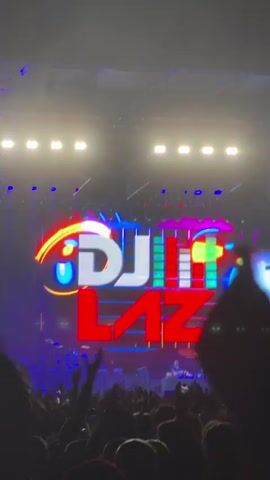 The video opens on a silhouetted crowd with their hands raised in the air. A large LED screen behind them displays the text DJ LAZ in large, stylized letters. The letters are outlined in bright blue and pink, with a rainbow of colors filling the gaps in the design. The word DJ is in a lighter gray, while LAZ is in a vibrant red. Above the letters, a pattern of colored bars fluctuates, resembling an equalizer. The stage is dimly lit, with a few spotlights illuminating the performers at the DJ booth. The overall atmosphere is that of a live concert or music festival. As the video progresses, the lighting on the screen shifts, with the blue outlines around the DJ LAZ text pulsing and changing color. The crowd remains engaged, with many individuals visible in silhouette. The focus remains on the screen and the energetic presentation of the DJ's name.