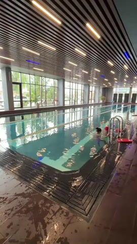 This video captures a scene inside an indoor swimming pool. The pool is long and divided into lanes, indicated by white lines on the bottom. The water is a clear turquoise color, and lights are visible beneath the surface, casting a blue glow. The pool area is surrounded by a dark, wet tiled floor, with a metal grate system along the edge.

The ceiling is a prominent feature, constructed from wooden slats arranged in a curved, modern design. Embedded within the ceiling are rows of bright, linear lights, with some additional blue lighting elements scattered throughout. Large windows line one side of the pool, offering a view of the outdoors, with trees and buildings visible beyond.

In the water, there are two individuals. One appears to be an adult female, possibly an instructor, wearing a light-colored swimsuit. She is positioned near the edge of the pool, holding onto a metal ladder. The other individual is a child, wearing a red swim cap and floatation devices around their arms. The child is also near the ladder, interacting with the adult. A red kickboard rests on the pool edge nearby. Several large, dark rectangular objects are lined up on the far side of the pool, possibly speakers or equipment. The overall impression is of a clean, well-maintained, and modern swimming facility.