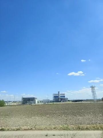 A wide shot shows a clear blue sky with a few white clouds. In the distance, there are industrial buildings and structures. A patch of dry, brown earth occupies the foreground and middle ground. There are no people or discernible actions visible in the frame. The scene appears to be an open, undeveloped area near industrial facilities.