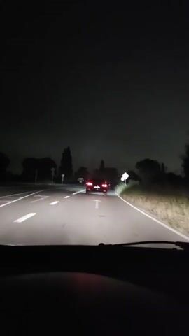 A first-person perspective video from inside a vehicle at night shows the vehicle driving on a two-lane road, following another car. The lead car is dark colored with its brake lights illuminated. The road is paved with white lane markings. There are trees and bushes on the sides of the road. Two road signs are visible, one a white circular sign with a red border, and another a yellow diamond-shaped sign with a black border. The sky is dark and appears overcast. The video zooms in on the rear of the car ahead, showing its license plate which reads "B 2333 KP". The lead car appears to be an SUV or similar vehicle.
