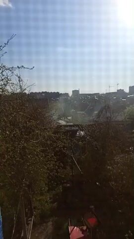 The video shows a view overlooking a landscape with various elements. In the foreground, there are trees or bushes with green leaves, along with a few objects including what appears to be a red table and some other equipment. In the middle ground, there are rooftops of houses, and some smoke or mist appears to be rising into the air. In the background, a skyline of buildings is visible, with some taller structures and construction cranes standing out against the clear sky. The sky is light blue with the sun shining.