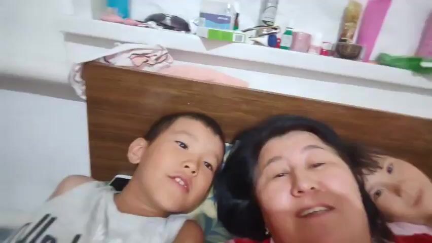 A family is taking a selfie in bed. A woman with dark hair and a red shirt is in the foreground, smiling. Next to her is a young boy with short dark hair and a grey tank top, also smiling. To the right of the woman, a young girl with dark hair and a pink shirt is visible, smiling. They are lying in bed with a wooden headboard behind them. A shelf with various items is visible above the headboard. The overall mood appears to be warm and familial.