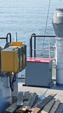 A video clip shows a section of a ship's deck with water in the background. In the foreground, there is a pile of wooden planks, some of which are weathered and splintered. To the left, a gray storage container with a red lid is visible. Behind the container, a railing with a chain is present. To the right, a large, cylindrical white structure stands, topped with a grey, conical object that has a label on it. Adjacent to this structure, an orange life preserver with white stripes is hanging on a railing. Further to the right, a small yellow box-like object is mounted on a metal support. The water appears choppy, indicating the ship is in motion. The overall scene suggests a functional, working deck of a vessel at sea.