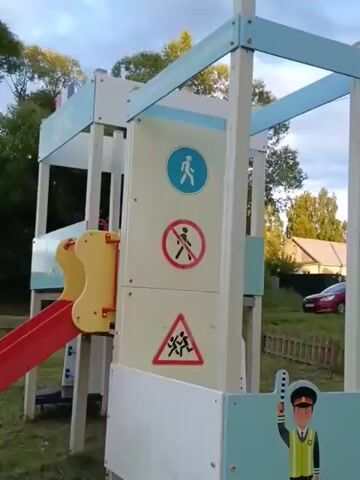 The media file shows a playground scene in Smolino, Russia. The playground features a red slide, a white and blue climbing structure with various safety signs, including a pedestrian crossing symbol, a "no walking" symbol, and a triangle with a running child silhouette. There is also a cartoon depiction of a traffic controller on the lower part of the structure.

In the foreground, a woman in a tan tracksuit and pink shoes is holding the hands of a young child wearing a blue shirt and a grey hat. The child is standing at the bottom of the slide. The woman appears to be assisting the child. The overall mood seems to be a calm, outdoor activity.

The background shows a residential area with houses and fences, along with trees and greenery, suggesting a suburban or rural setting. A red car is visible driving on a road in the distance. The sky is overcast, indicating it might be evening or a cloudy day. There is no visible text on the playground structures other than symbols.
