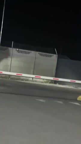 The media file shows a dark, outdoor scene at night. The primary subject is a large, blue semi-truck with its headlights on, parked or moving slowly near a barrier gate. The barrier gate is white with red markings and appears to be partially lowered. In the foreground, there is a paved road with white arrow markings.

Behind the truck and barrier, a concrete fence or wall is visible. There are traffic signs present: a yield sign (red triangle with white center) and a directional sign with arrows. A street lamp is also visible. The ground appears to be covered with grass and dirt near the fence.

The overall atmosphere is that of a nighttime security checkpoint or entrance, possibly at a facility or industrial area. The lighting is artificial, provided by the truck's headlights, the street lamp, and possibly interior lights within the truck cab, which show a faint blue glow. The time of day is clearly night due to the dark sky and artificial lighting. There are no people visible in the media. The location cues, such as the style of the fence and traffic signs, are consistent with infrastructure found in various countries, but the prompt specifies the location as Nerubais'ke, Ukraine. There is no visible text in the media file itself, other than markings on the traffic signs.