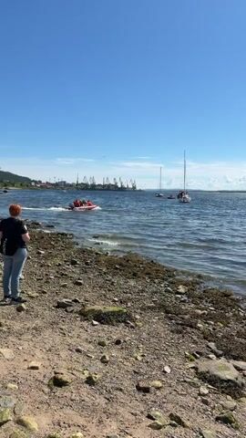 A bright, sunny day with a clear blue sky over a body of water. In the foreground, a rocky, pebbly shore is visible. To the left, a person with reddish hair stands facing the water, holding a phone. Further out in the water, a red inflatable boat with several people on board moves from left to right. A white sailboat is anchored to the right of the inflatable boat. In the background, a shoreline with buildings and industrial cranes is visible. The water is calm with small waves.