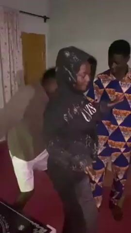 Three young people are dancing in a room with red flooring. One person is wearing a black hooded sweatshirt with the word "STATUS" written on it and is mostly facing forward. A person to their left is wearing a khaki shirt and white shorts, smiling and bobbing. Another person to the right is wearing a blue and orange patterned shirt and pants, and is moving their arms slightly. The room also contains a small table and a window with curtains. The lighting in the room is dim.
