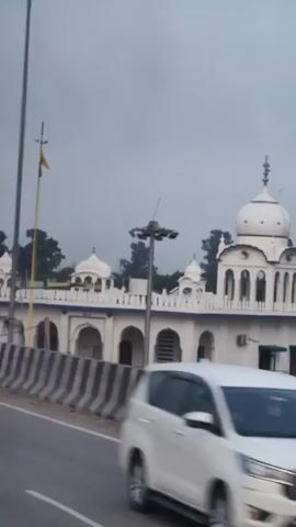 The video shows a white Sikh gurdwara, a religious building for Sikhs. The gurdwara has a large central dome and several smaller domes, as well as arched windows and a decorative blue trim. The building is white with white domes. It is daytime and the sky is overcast. The camera appears to be mounted on a moving vehicle, as indicated by the motion blur and the presence of a highway barrier in the foreground. The vehicle is moving past the gurdwara, providing a side view of the building.
