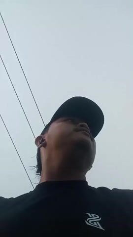 The video is a close-up, low-angle shot of a person wearing a black t-shirt and a black baseball cap. The person is looking upwards. The background is an overcast sky with several thin, dark lines that appear to be wires. The person's face is partially obscured by the brim of the cap. There is a logo on the t-shirt. The video is short and seems to be a brief clip.