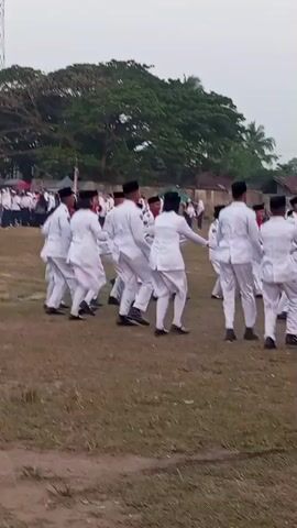 A group of people wearing white uniforms and black caps are dancing in an open grassy field. The dancers are moving in unison with synchronized steps and arm movements. The background shows trees, a fence, and some buildings. The sky is overcast.
