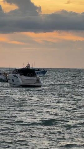 The video captures a tranquil scene of two boats sailing on a body of water during what appears to be sunset. The sky is adorned with warm hues of orange and yellow, interspersed with dramatic dark clouds, suggesting a dynamic weather condition. The sea is moderately rough, with visible ripples and waves, indicating a gentle breeze.

In the foreground, a white yacht with the name "SUNDANCE" clearly visible on its stern is anchored or moving slowly. Its sleek design suggests a modern vessel. Further in the distance, another boat, a narrower speed boat, is in motion, leaving a visible wake as it cuts through the water. The speed boat appears to be white with blue accents.

The overall mood is serene and picturesque, with the play of light and shadow on the water and in the sky creating a visually appealing contrast. The time of day is evident from the soft, golden light illuminating the clouds and horizon. The location is identified as Baan Bang Rak, Thailand. The scene evokes a sense of calm and perhaps the end of a day of leisure on the water. There are no people clearly visible in the video, focusing the attention on the vessels and the natural environment.