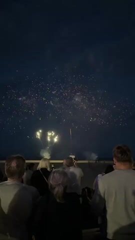 The video shows a crowd of people watching fireworks display at night. The sky is dark and filled with smoke and bursts of light from the fireworks. The people are silhouetted against the bright sky, and they appear to be standing on a beach or a similar outdoor venue. The fireworks are colorful, with explosions of red, yellow, and white lights. Some of the fireworks are large and elaborate, while others are smaller and more scattered. The crowd is diverse, with people of all ages and backgrounds. They are all looking up at the sky, mesmerized by the spectacle. The atmosphere is one of excitement and awe.