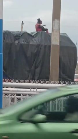 The image depicts an unusual outdoor scene, likely on a street or highway in Kawo, Nigeria, during what appears to be an overcast daytime.

In the mid-ground, a person is seated on top of a large vehicle, which is heavily covered by a black tarpaulin. The vehicle's obscured shape suggests it could be a truck or lorry. The individual is dressed in a red top and dark trousers, with their face completely obscured by a dark mask or balaclava. They are holding what appears to be a stringed musical instrument, possibly a guitar or a similar local instrument.

In the immediate foreground, a white, ornate metal railing, likely part of a bridge or overpass barrier, is visible, suggesting the photograph was taken from an elevated position. A blurry light green car occupies the very bottom of the frame, indicating ongoing traffic. To the right, a tall, light brown utility pole stands, with some faded, unreadable text visible on its surface. In the background, behind the pole, a faint structure resembling scaffolding or an incomplete building can be seen. The overall scene captures an unconventional event in an urban setting.