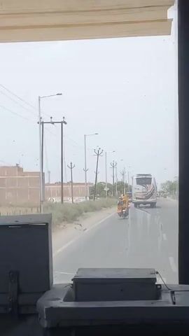 A view from inside a bus driving down a paved road with power poles on either side. A person on a scooter wearing a yellow garment is on the road ahead of the bus, in the same lane. A white bus is in the oncoming lane. There is a building on the left side of the road. The day is overcast. The dashboard of the bus is visible.