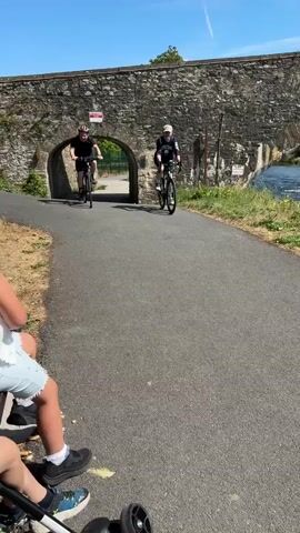 The video begins showing a paved path next to a grassy area. A stone archway spans across the path in the background. Two individuals on bicycles are approaching the archway from the opposite direction. In the foreground, two children and an adult are sitting on a bench, facing away from the camera. One of the children is wearing a yellow hat and a white shirt. A stroller is parked next to the bench. The cyclists pass through the archway and continue down the path. The video is shot on a sunny day with a clear blue sky. The environment appears to be outdoors, possibly a park or a recreational trail.