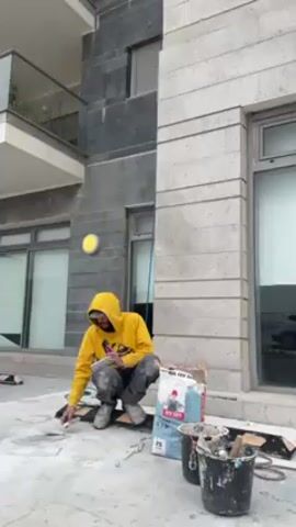 A man in a yellow hooded sweatshirt and gray cargo pants stands near a building, holding a smartphone. He squats down with his legs crossed at the ankles, then stands up. A bag of tile adhesive and buckets are on the ground near him. The building has windows and grey stone cladding. A blue rope hangs down the building's side.